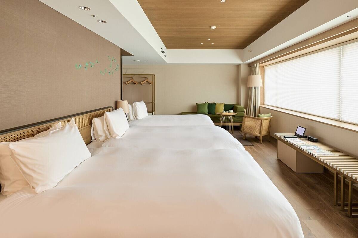 Ein Hotelzimmer mit einem großen Bett, einem Schreibtisch mit einem Laptop, einem Sessel, einem kleinen Tisch und einem Fenster mit Jalousien.