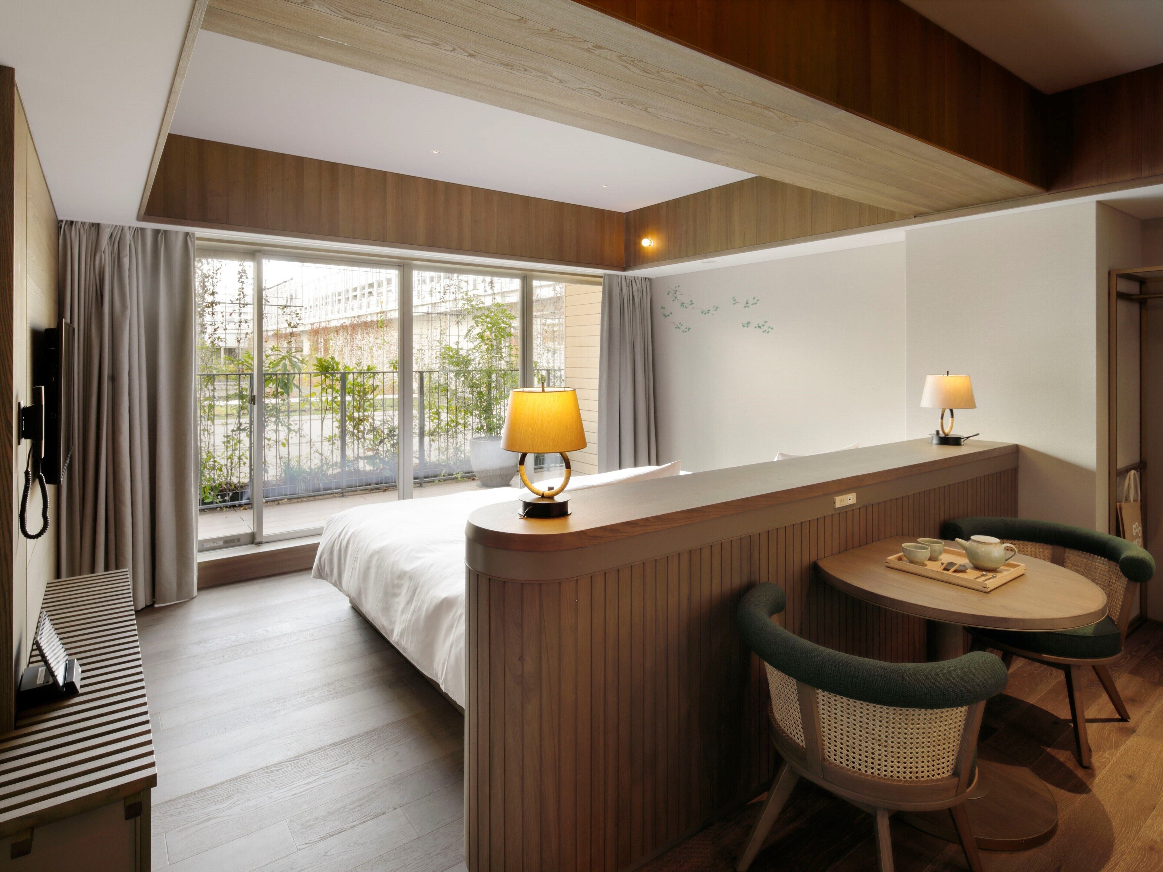 A hotel room with a bed, a desk with a lamp, a table with chairs, and a view of greenery outside.