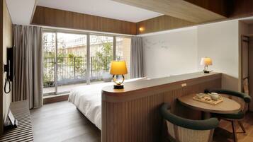 A hotel room with a bed, a desk with a lamp, a table with chairs, and a view of greenery outside.