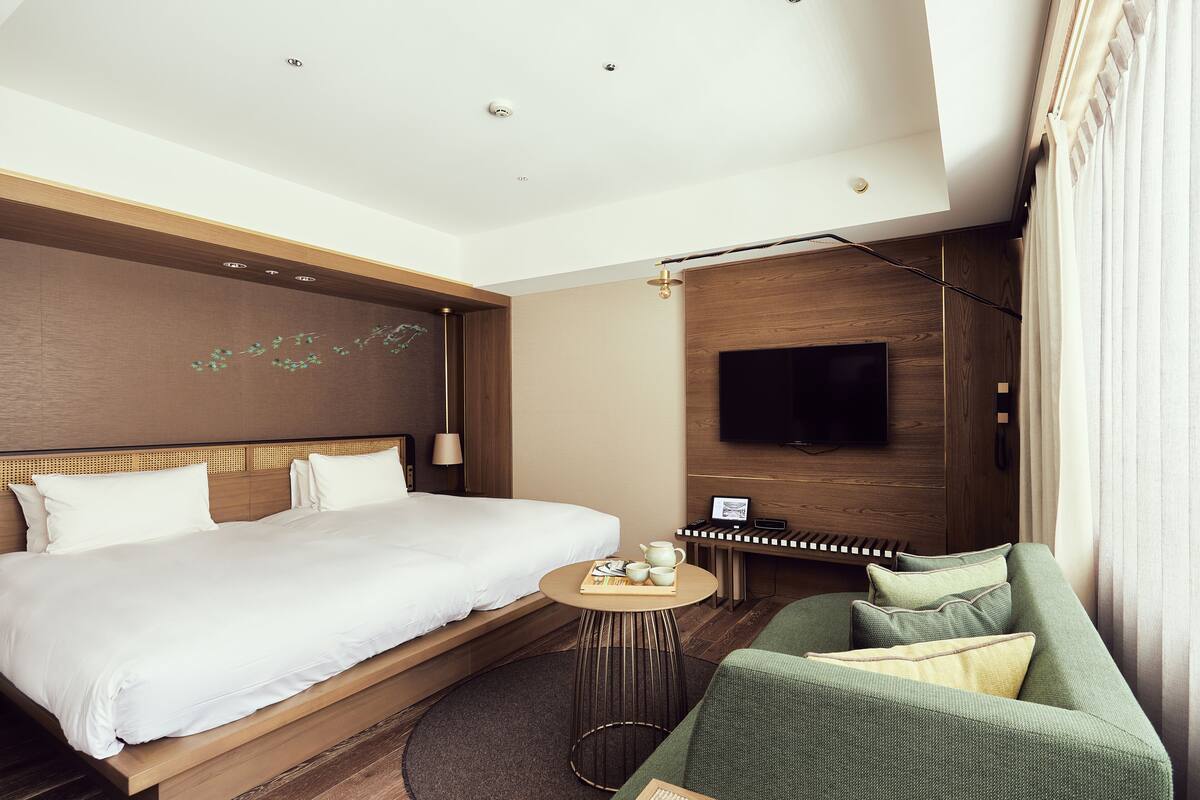 Ein modernes Hotelzimmer mit einem großen Bett, einem kleinen runden Tisch mit einem Buch und einem Teekessel, einer grünen Couch, einem an der Wand montierten Fernseher und einer Holzwand.