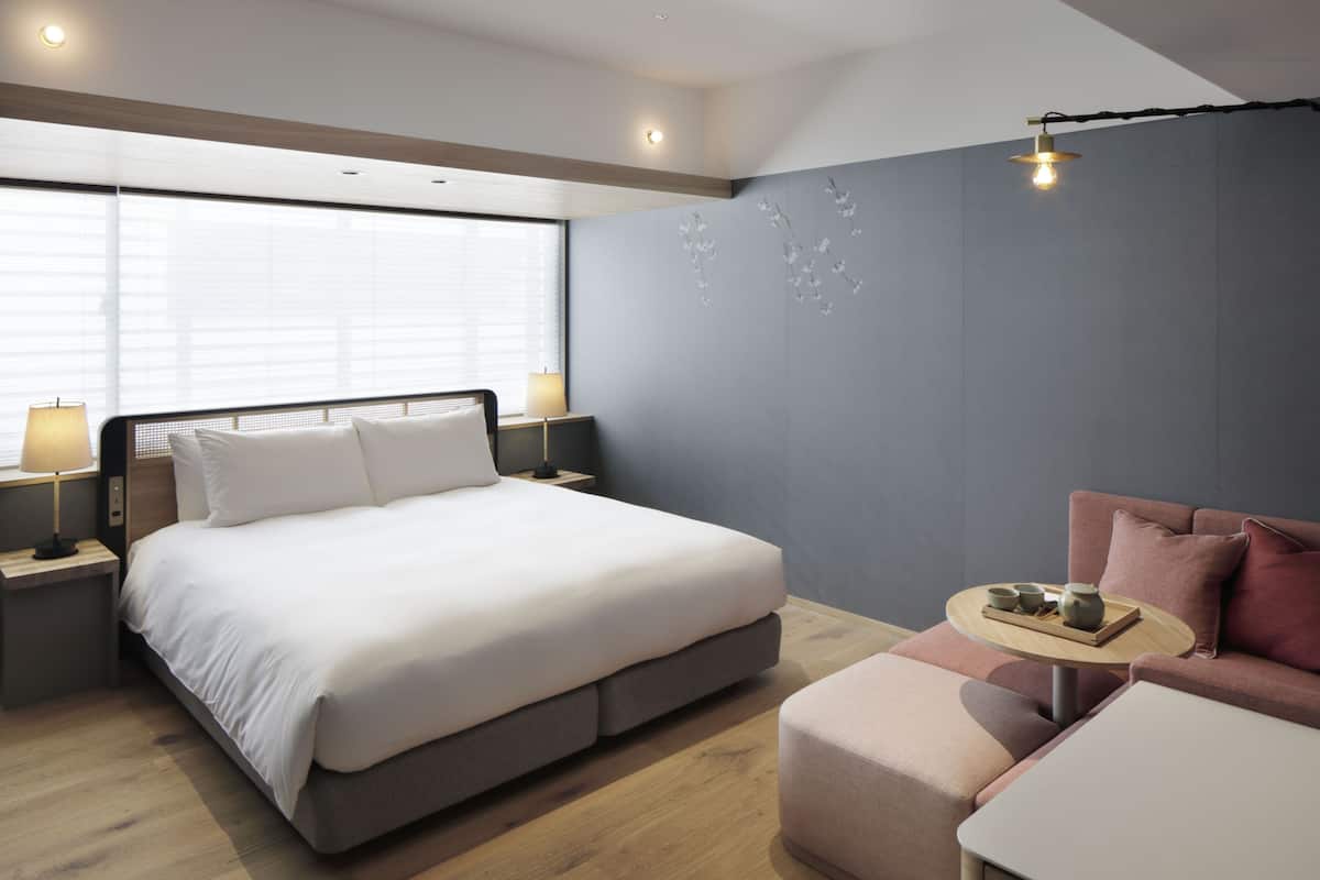 Ein modernes Hotelzimmer mit einem großen Bett, einer Sitzecke, einem kleinen Tischchen mit Tablett und einem Fenster mit Jalousien.