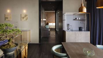 A modern hotel room with a wooden dining table, a kitchen area with a refrigerator, and a living space with a bed and a staircase leading to the upper floor.