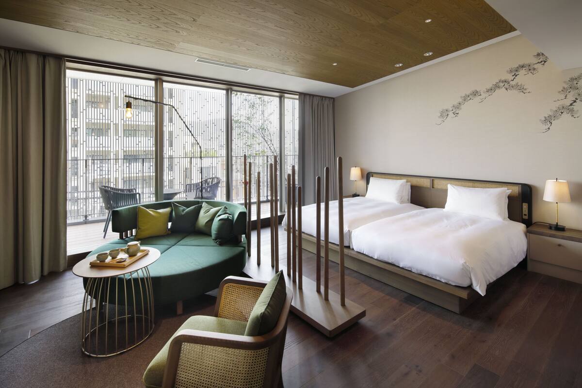 Ein modernes Hotelzimmer mit einem großen Bett, einem grünen Sofa, einem kleinen runden Tisch und Blick auf die Stadt durch große Fenster.