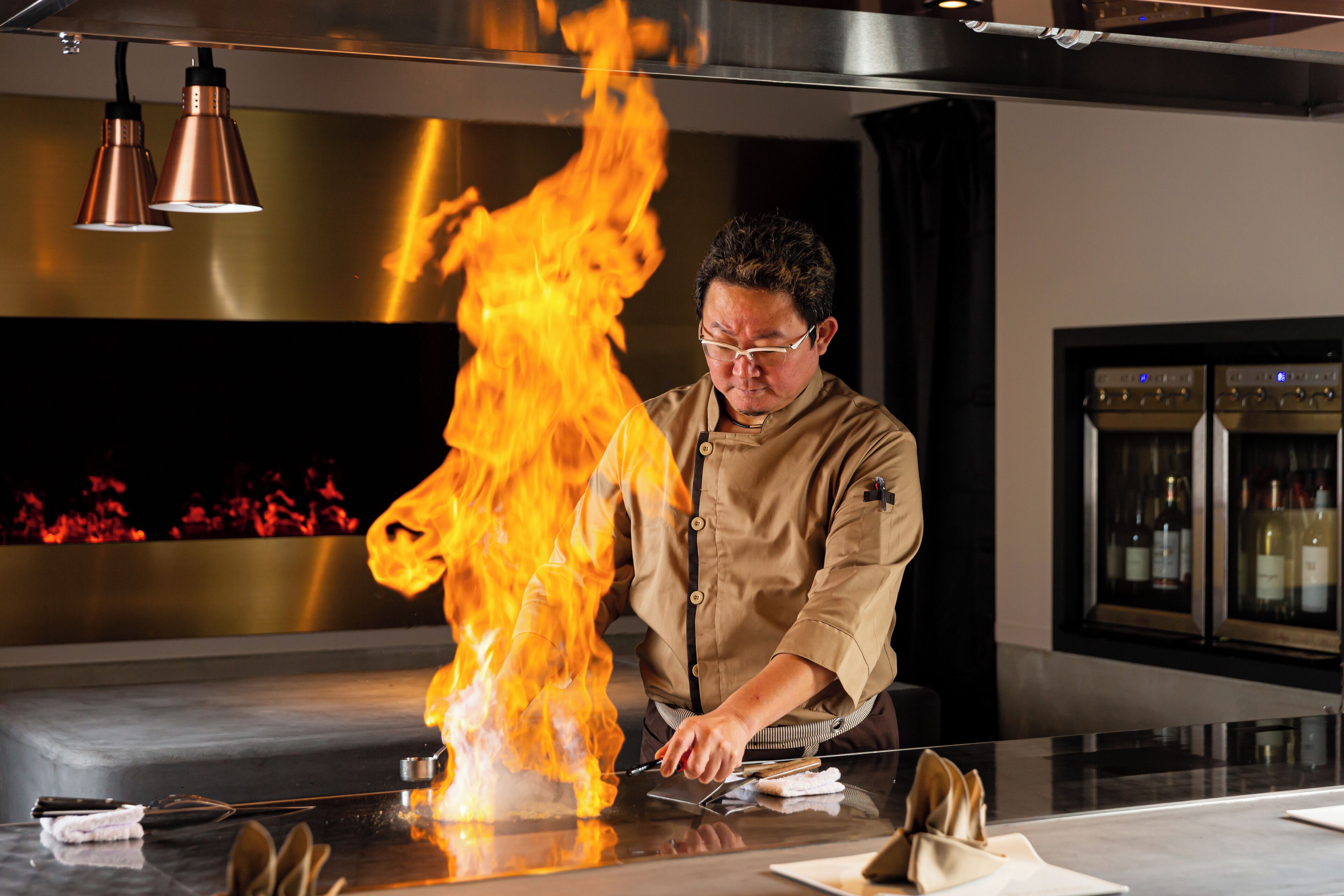 A chef performing a flambé with a large flame on a stainless steel table in a modern kitchen setting.