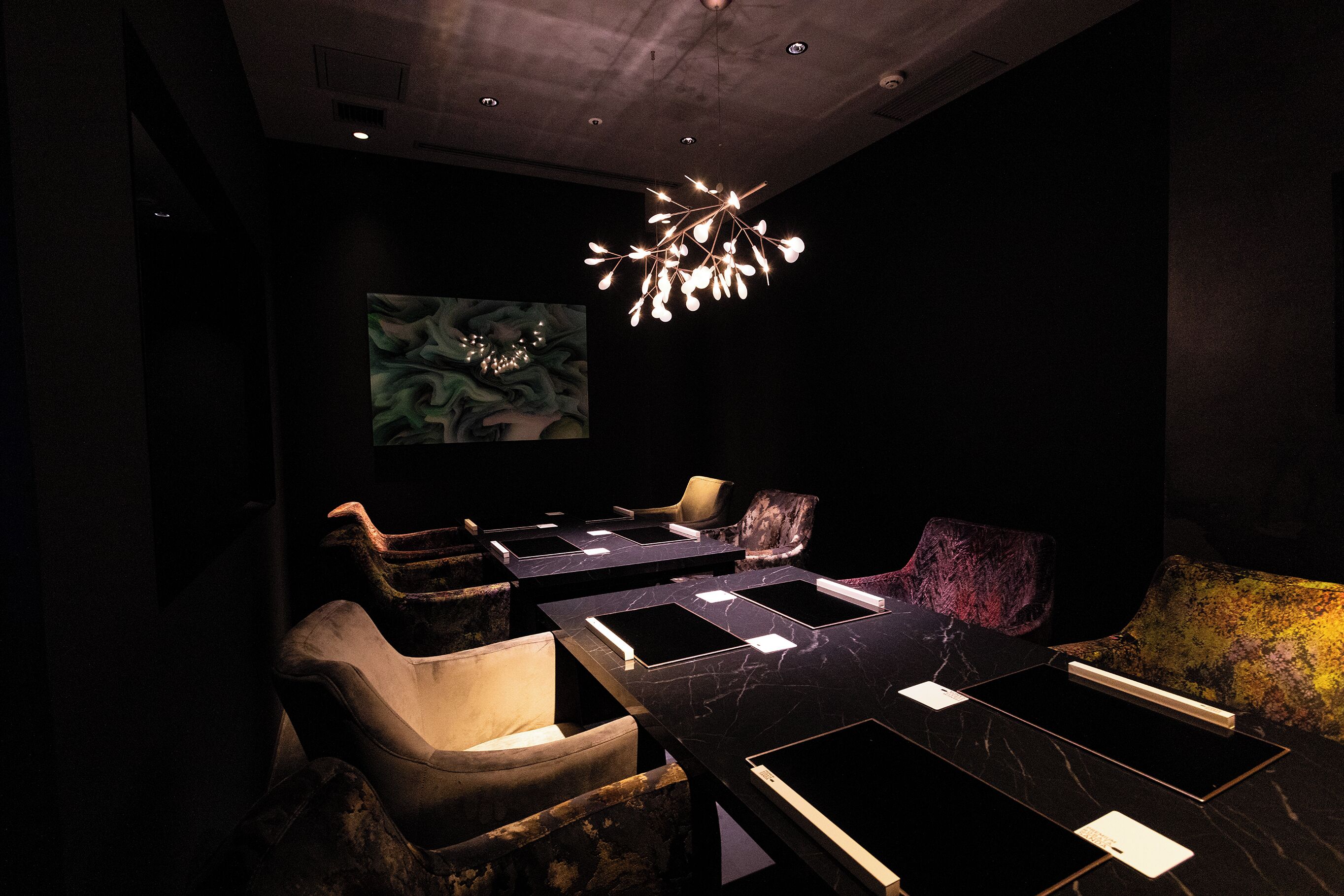 A dimly lit room with a modern chandelier, several leather chairs, and a large abstract painting on the wall.
