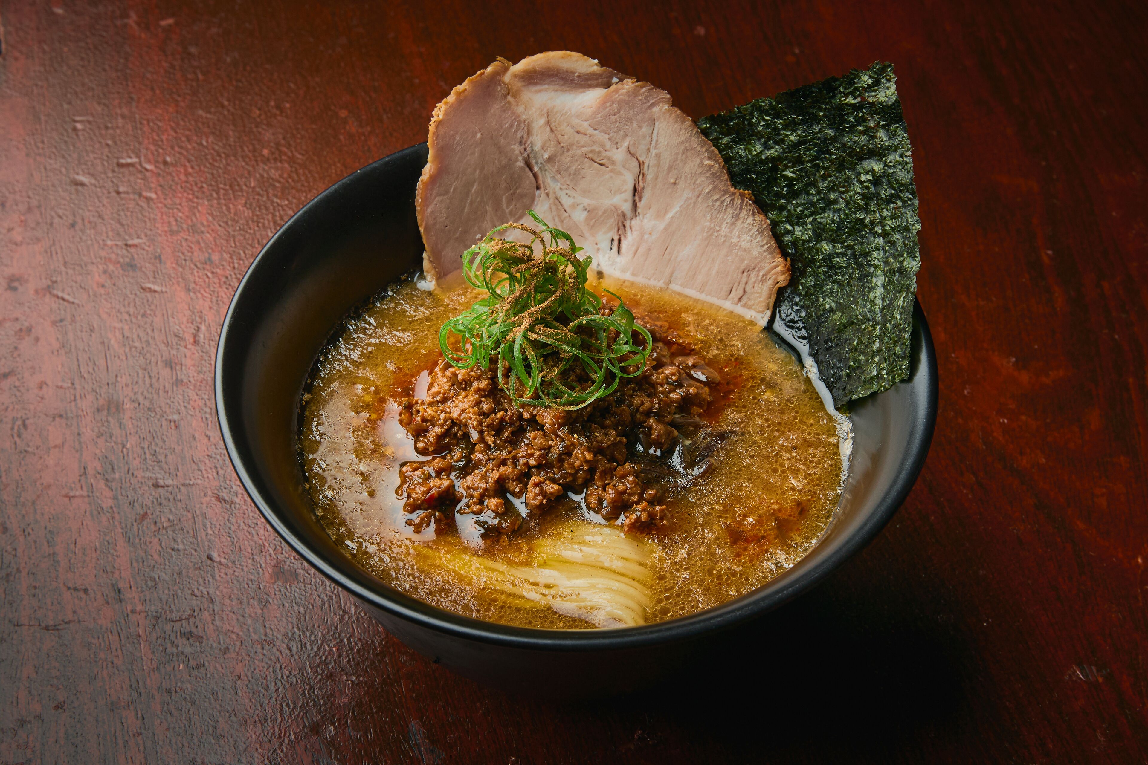 A bowl of soup with meat and vegetables, accompanied by a slice of meat and seaweed on a wooden table.