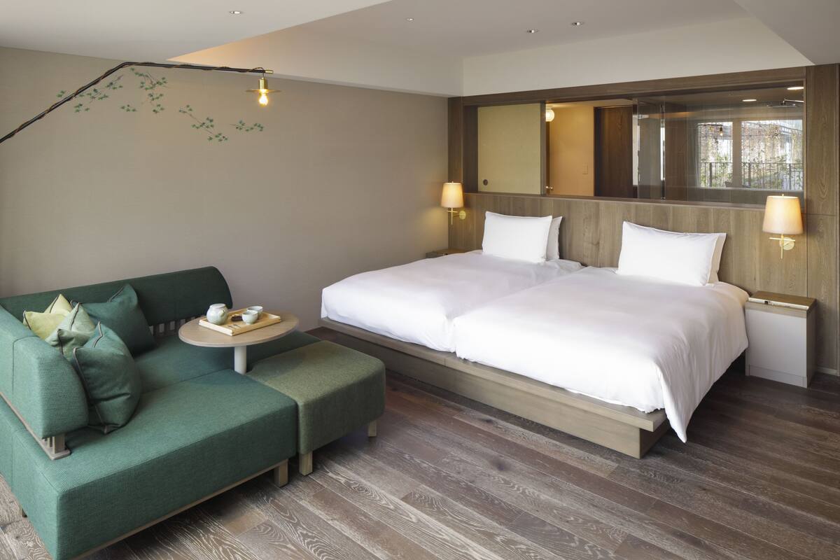 Ein modernes Hotelzimmer mit einem großen Bett, einem grünen Sofa, einem kleinen runden Tisch mit Tablett und einem großen Spiegel über dem Bett.