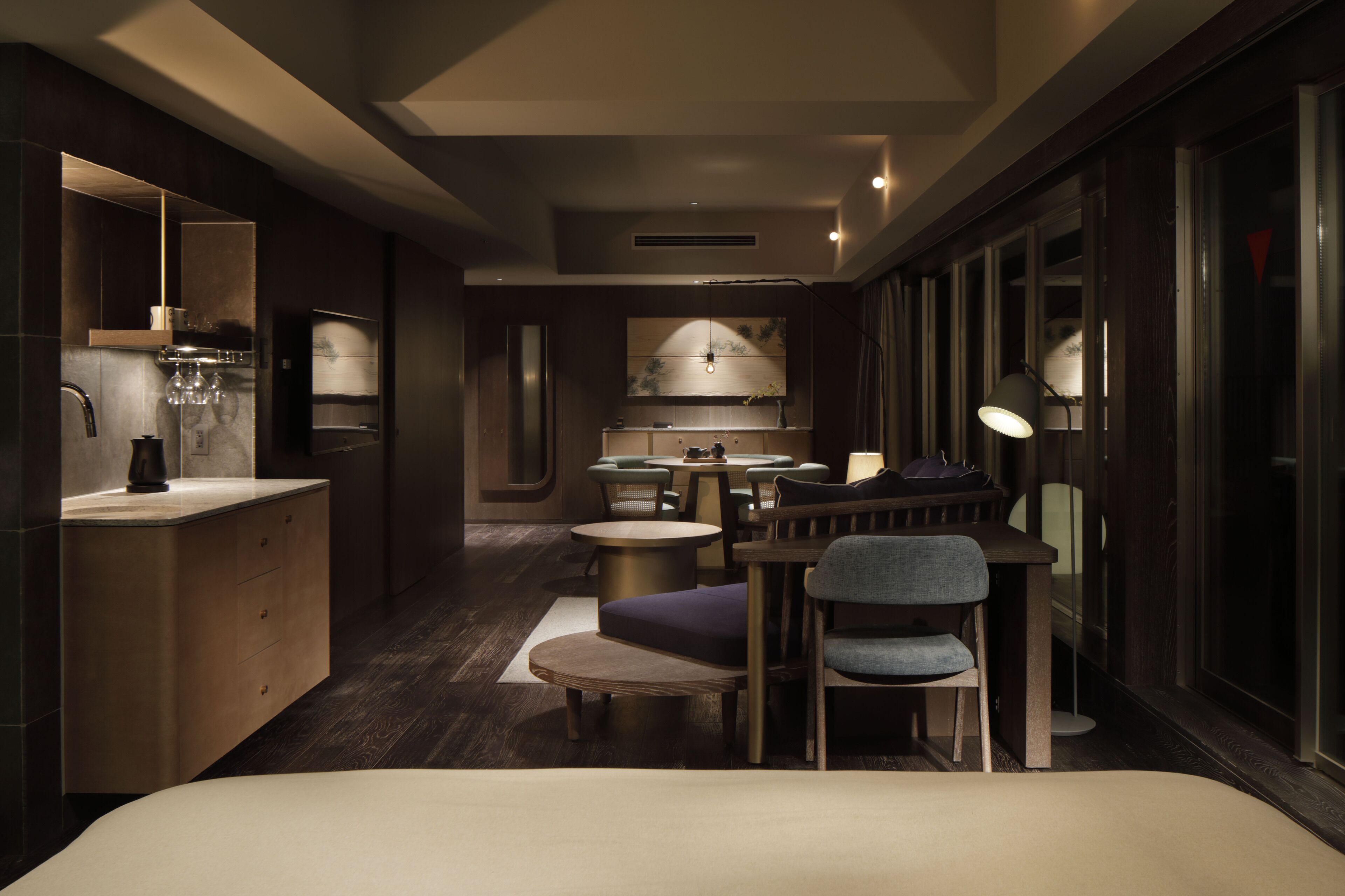 A modern hotel room with a bed, a small seating area, a kitchenette, and a large window.