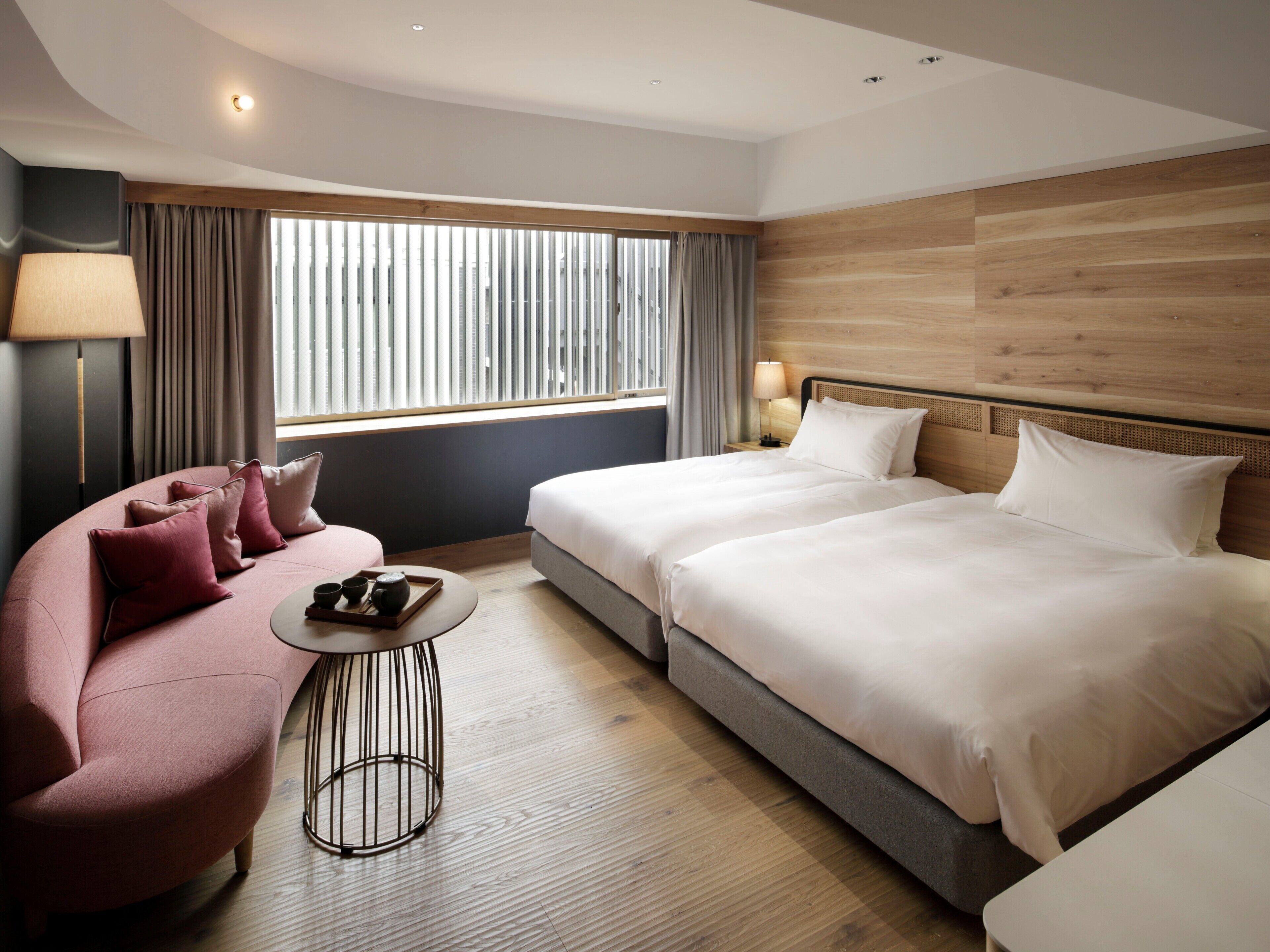 A modern hotel room with a large bed, a sofa, a small table with a tray of tea, and a window with blinds.