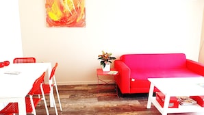 Ein Zimmer mit einem roten Sofa, einem weißen Tisch, Stühlen, einem kleinen Beistelltisch mit Pflanze und einem roten abstrakten Gemälde an der Wand.
