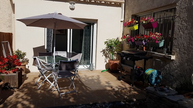 Eine Terrasse im Freien mit einem Glastisch und Stühlen, einem Grill und Blumentöpfen.