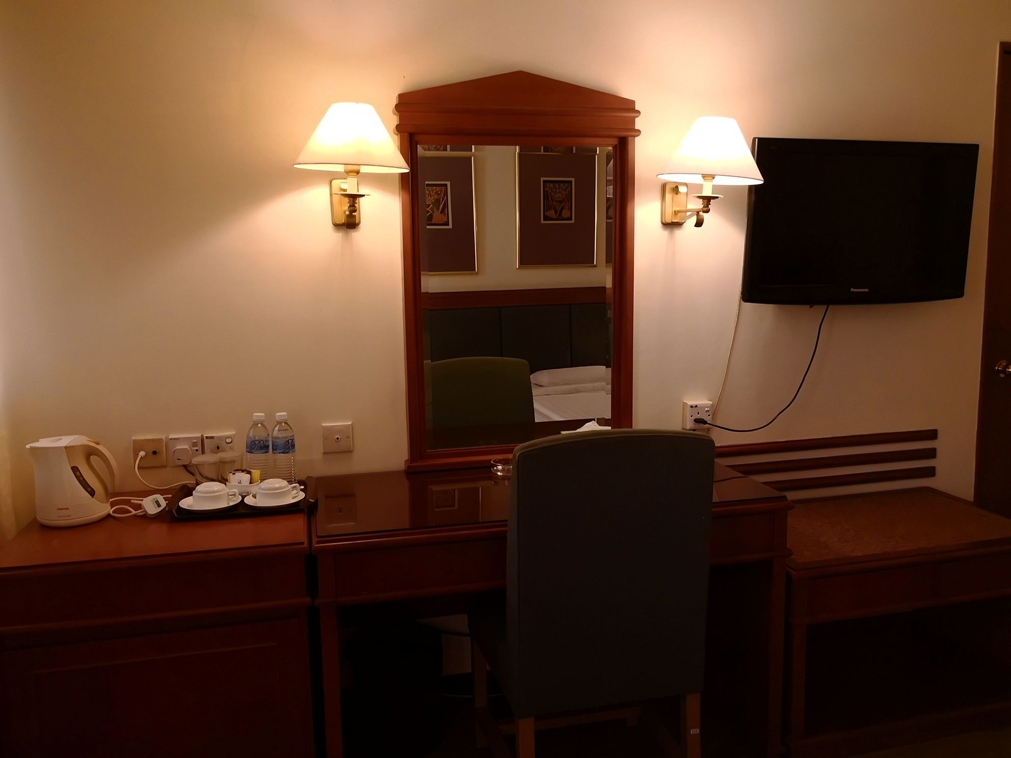 A hotel room with a desk, two bedside lamps, a flat-screen TV, a coffee maker, and a chair.
