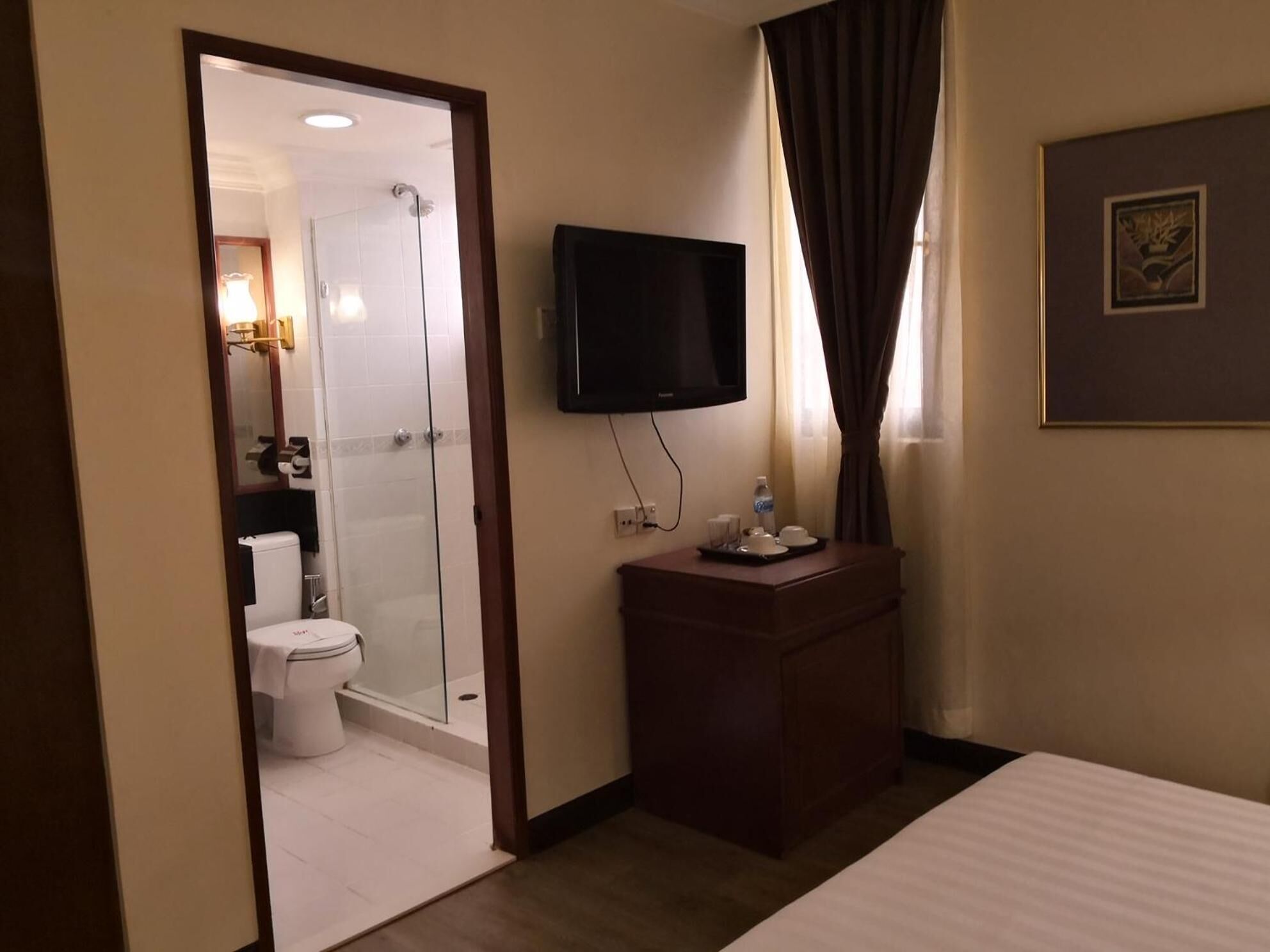 Habitación de hotel con cama, televisor montado en la pared, cómoda de madera, zona de ducha y baño con inodoro y lavamanos.