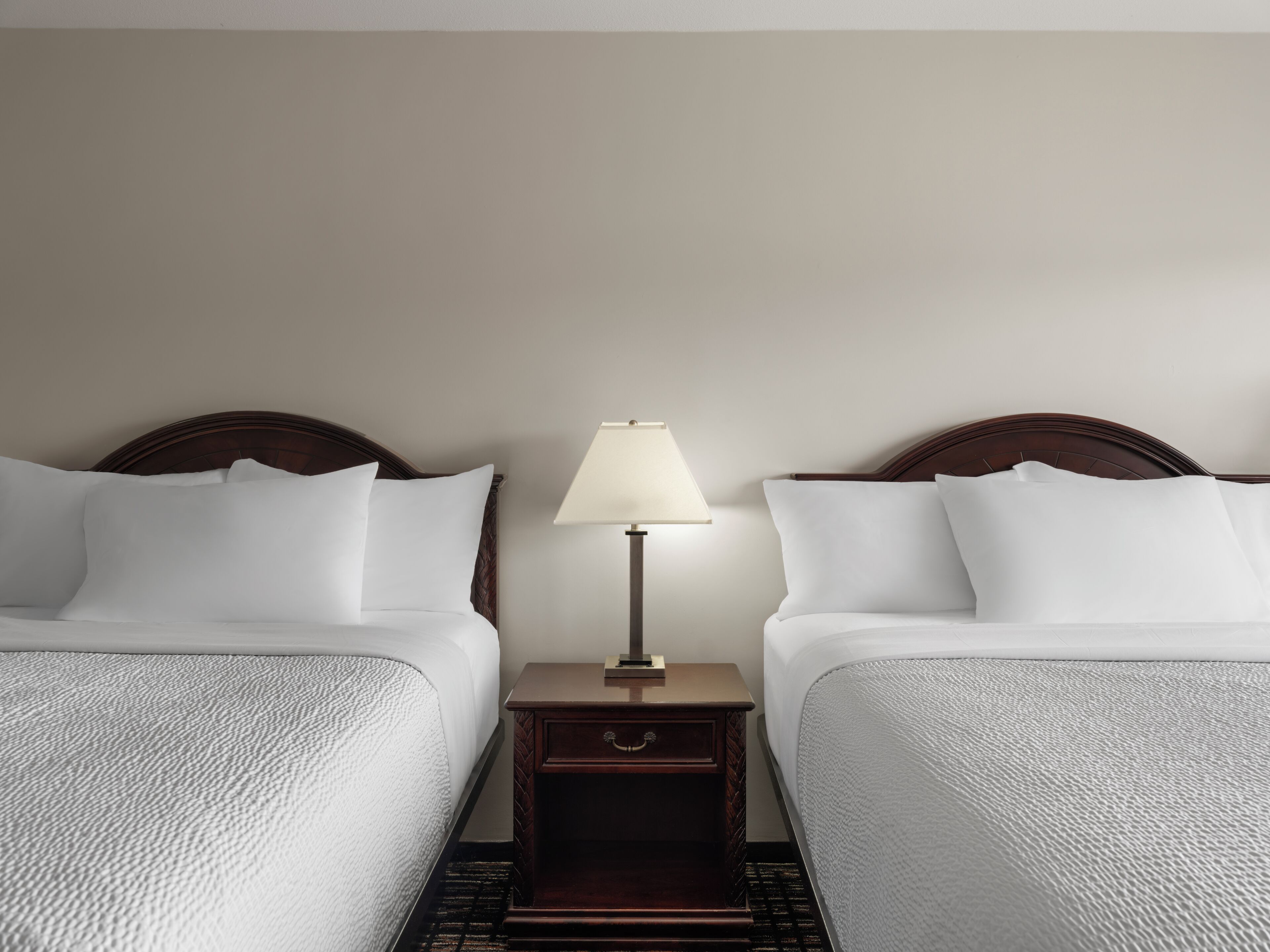 Ein Hotelzimmer mit zwei Betten, einem Nachttisch mit Lampe und einer schlichten Wand.