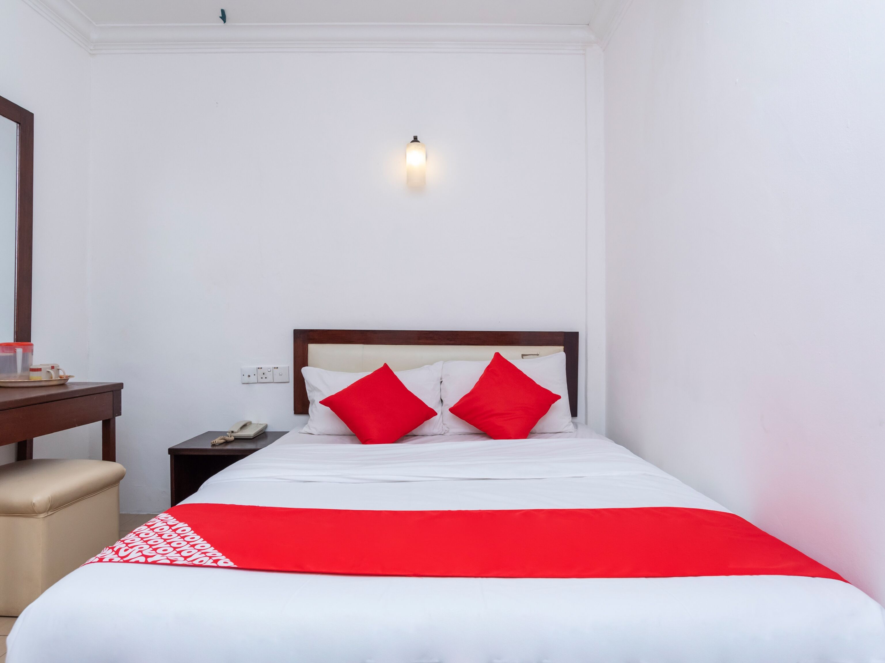 A neatly made bed with a red and white bedspread, two red pillows, and a wooden headboard. There is a small bedside table with a phone and a cup.