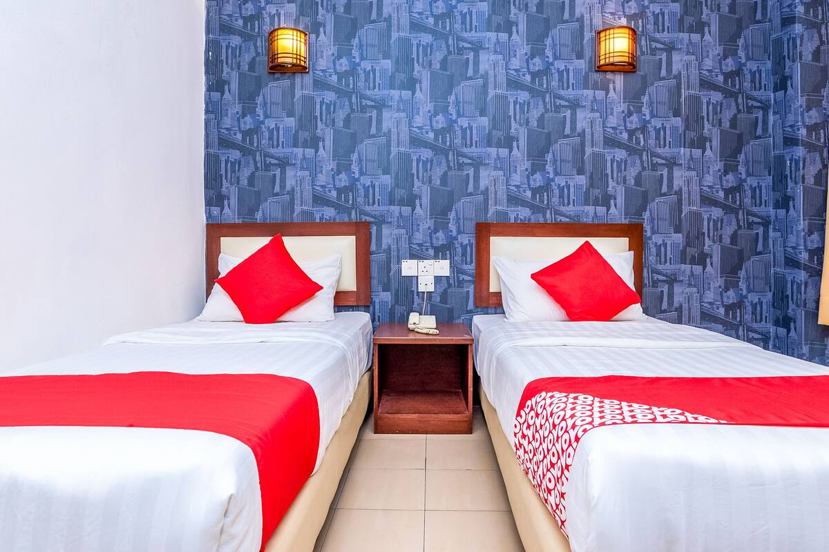 A hotel room with two beds, red and white bedding, a patterned blue wall, and two wall-mounted lamps.