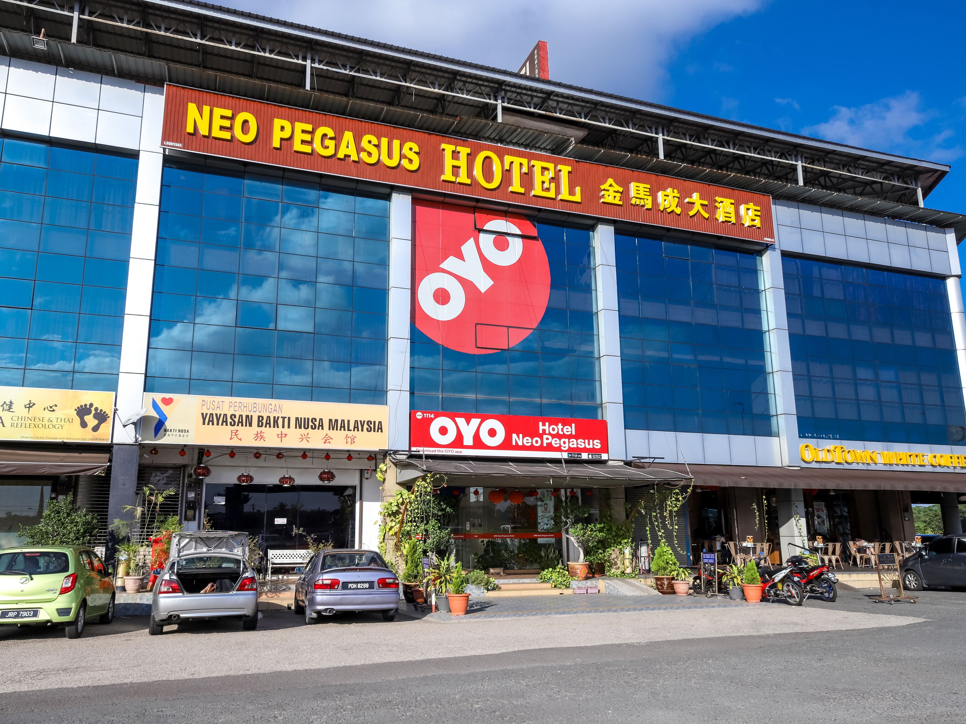 Hotel Neo Pegasus amb un gran cartell vermell i blanc, diversos cotxes aparcats al davant i plantes en testos al voltant de l'entrada.