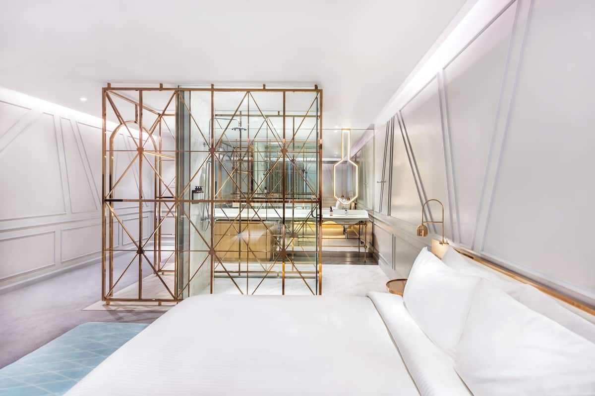 A modern hotel room with a large bed, a glass partition, and a bathroom with a mirror and sink.