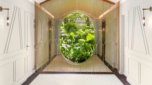 Un pasillo con un ventanal circular que muestra abundante vegetación, paredes blancas y un suelo de baldosas.