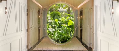 A hallway with a circular window revealing lush greenery, white walls, and a tiled floor.