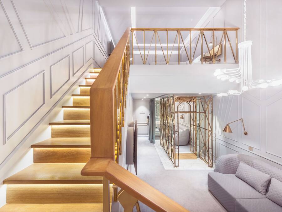 Una escalera moderna con escalones de madera y barandilla de metal, que conduce a una zona en el segundo piso con un separador de vidrio y una zona de estar acolchada.