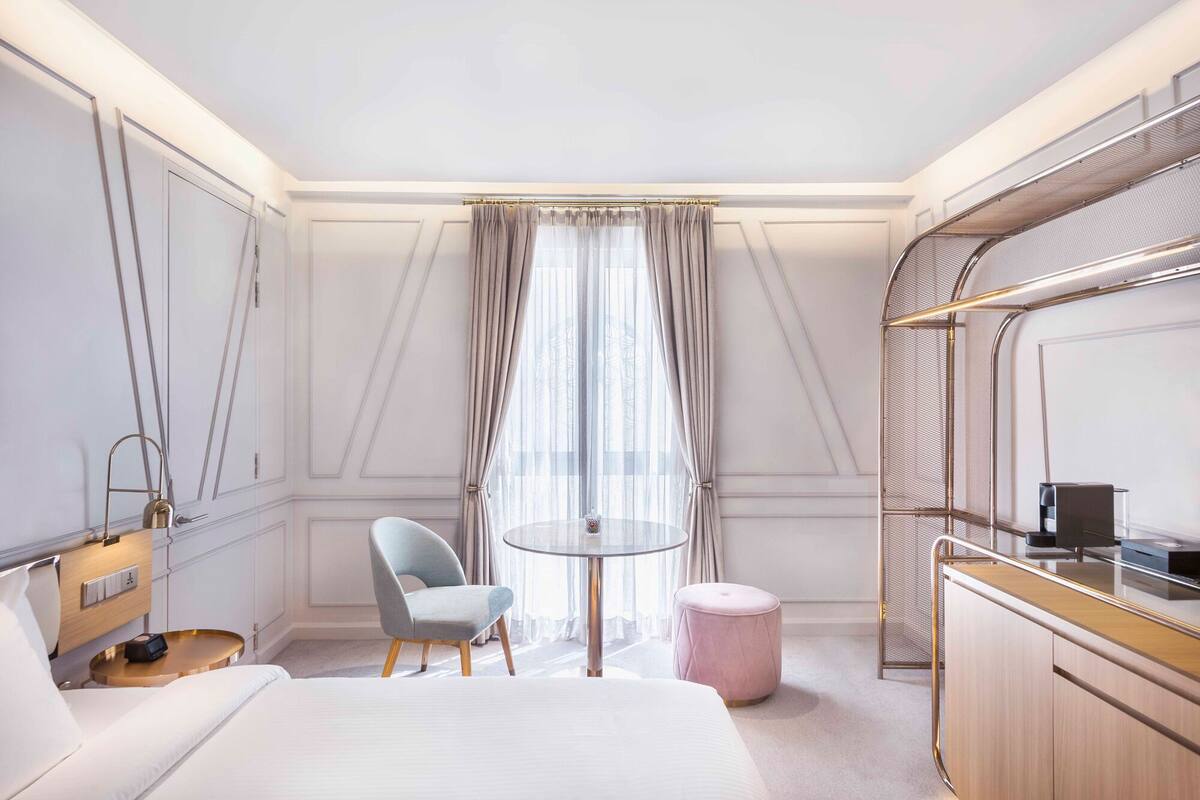 A hotel room with a bed, a round table, a chair, a small ottoman, a mirror, and a wardrobe.