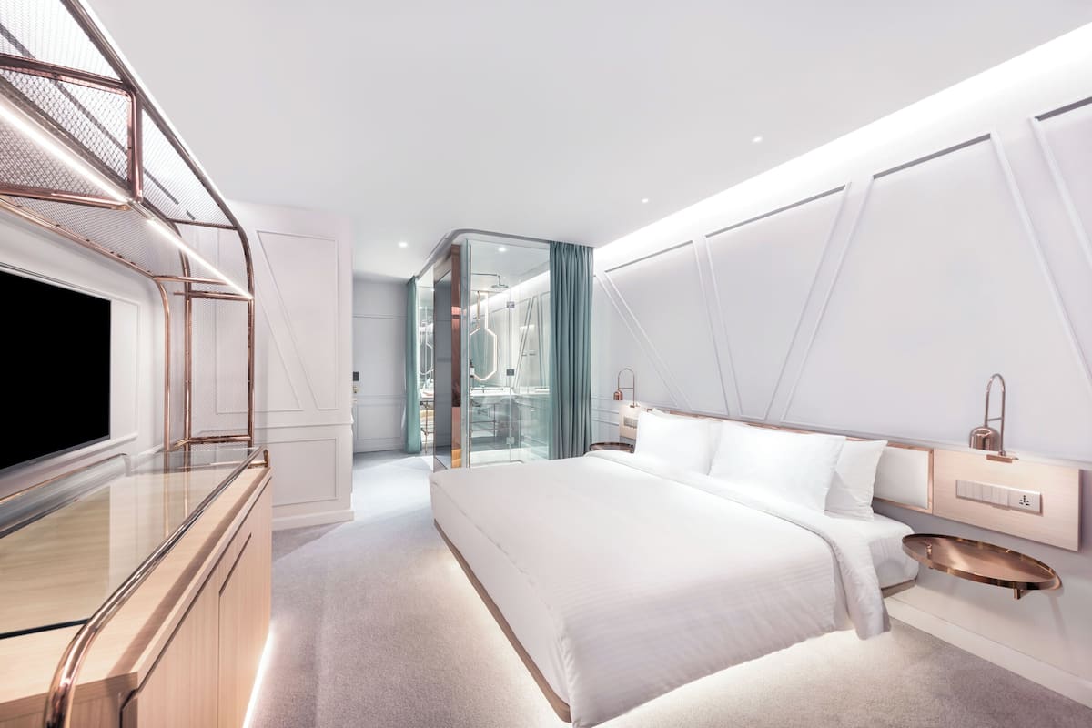 A modern hotel room with a large bed, a TV, a glass-enclosed bathroom, and a unique ceiling design.