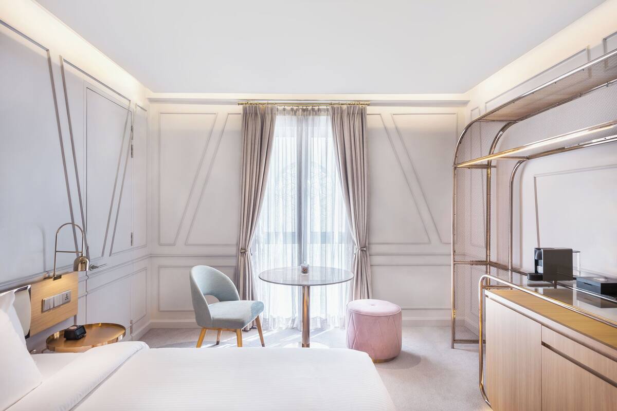 A hotel room with a bed, a round table, a chair, a small ottoman, and a wardrobe.