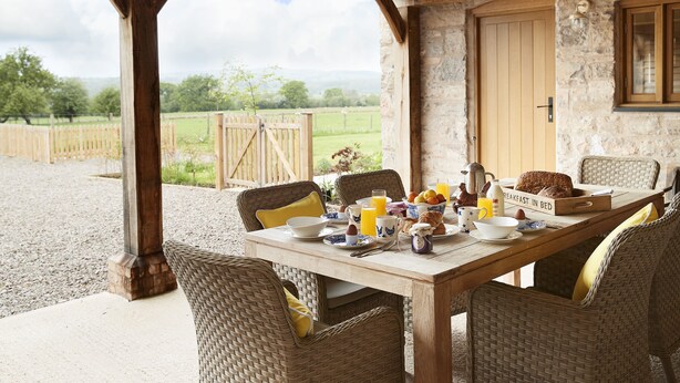 A rustic wooden table set for breakfast with wicker chairs, a stone wall, and a view of the countryside.