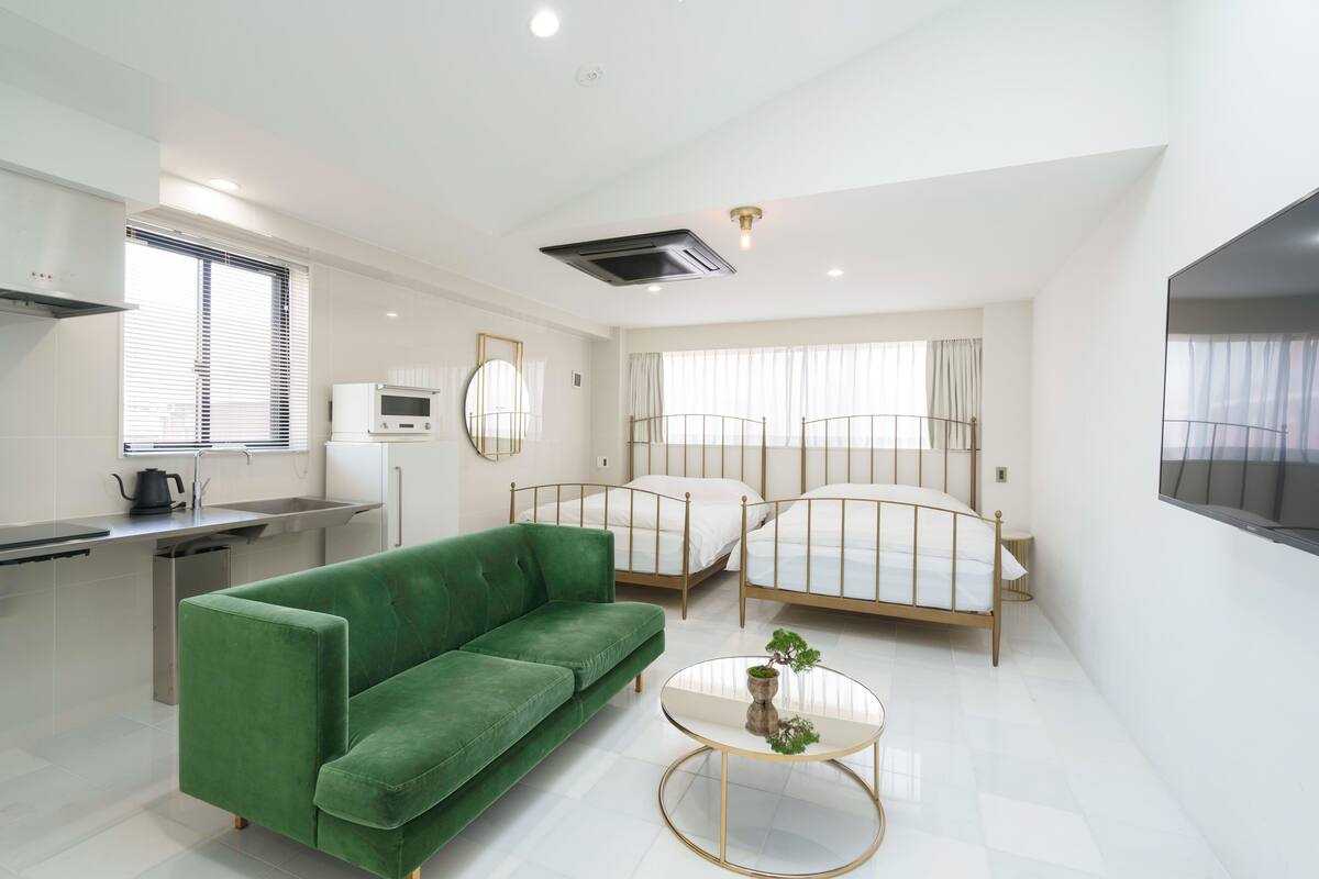 Une chambre d’hôtel moderne avec un canapé vert, deux lits, un petit coin cuisine et un téléviseur fixé au mur.