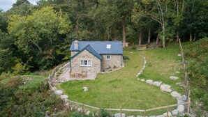 Une maison en pierre avec un toit bleu, entourée d’un mur de pierre et d’une cour verdoyante où se trouve un banc en bois.