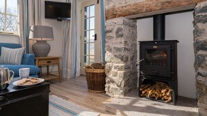 Un salon chaleureux avec une cheminée en pierre, un téléviseur fixé au mur, un canapé avec des coussins, une table basse sur laquelle se trouve une assiette de biscuits et un panier en osier.