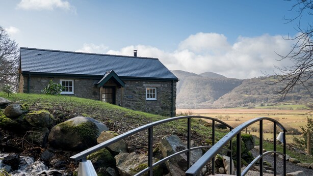 Une maison en pierre, reliée à l’extérieur par un pont métallique, et entourée d’un paysage montagneux.