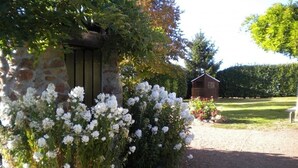 A stone wall with white flowers, a wooden structure, and a hedge in a garden setting. - Cottage 80m² with Terrace, Wifi and Pets Allowed (Notre-Dame-de-Boisset)