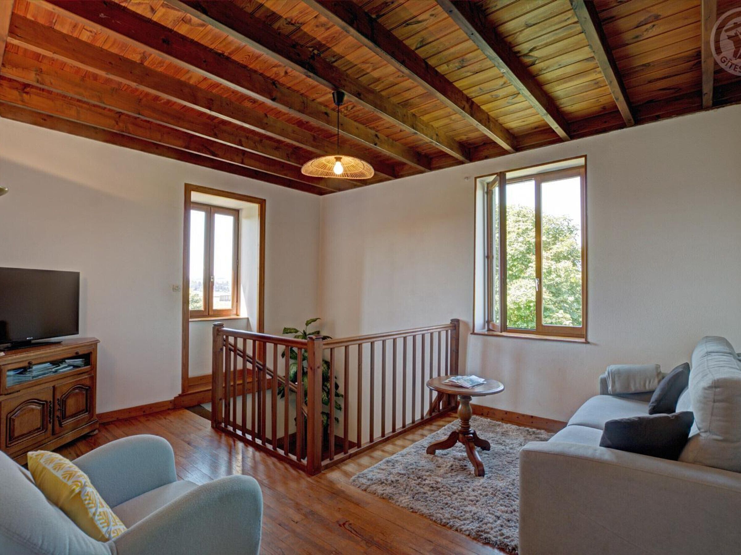 Ein geräumiges Wohnzimmer mit Holzbalken, einem Fernseher, einer Couch, einem Couchtisch und einer Treppe mit Geländer.
