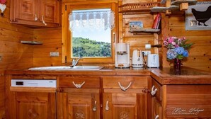 Cozinha de cabana de madeira com janela para uma paisagem verde, uma cafeteira, uma torradeira e uma máquina de lavar louça.