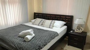 A neatly made bed with a dark grey blanket and two rolled towels, a bedside table with a lamp, and a small drawer. - Posada Nativa Nilvis Place (San Andrés)