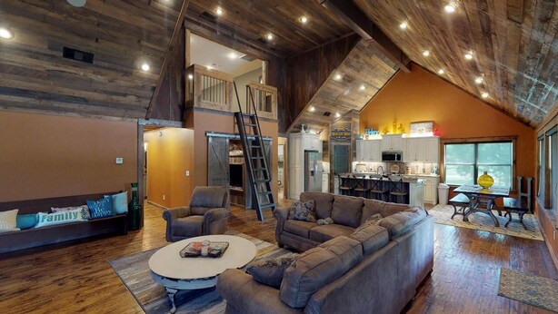 Un vaste espace de vie aménagé dans un style industriel, avec un plafond haut, des poutres en bois, un grand canapé, une table basse et une cuisine à l’arrière-plan.