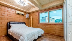 A bedroom with a wooden ceiling, brick walls, a bed with a white bedspread, a window with a view of buildings, and a ceiling fan. - Haedoji Hwangto Town (Jeju City)