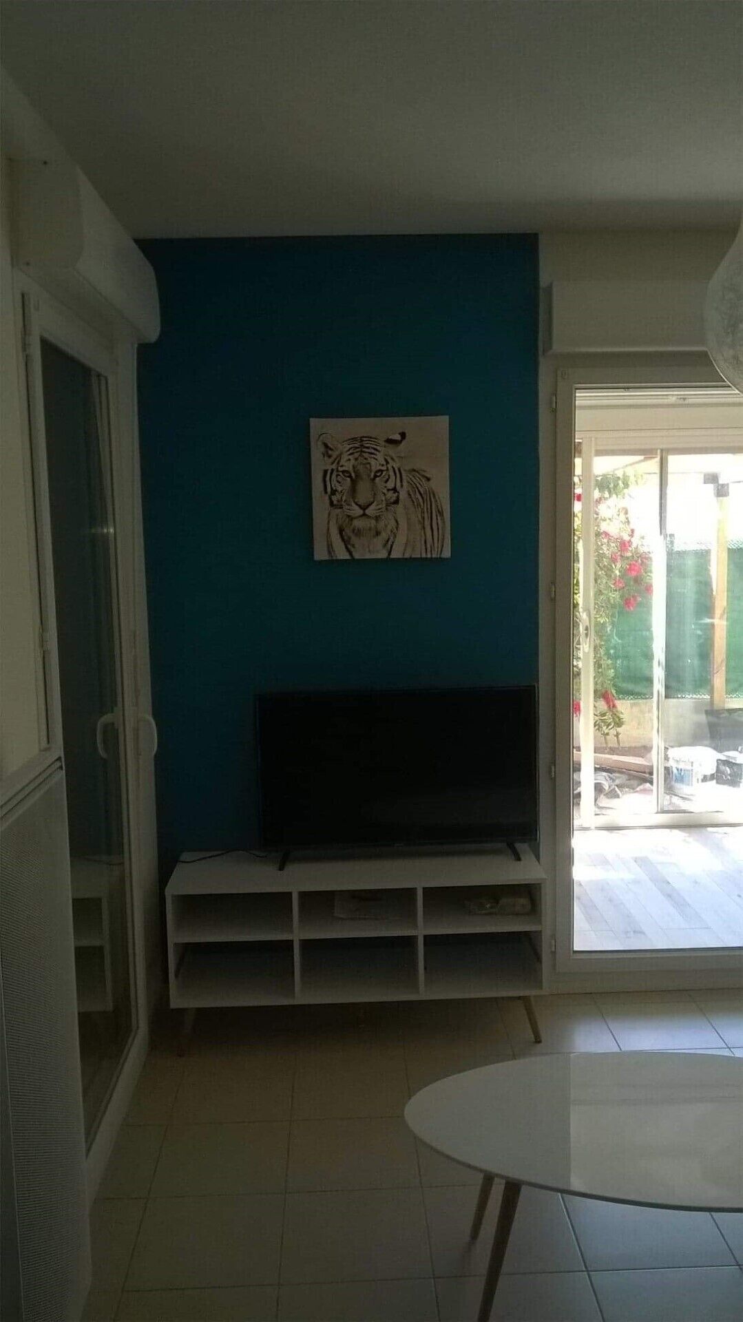 Ein Zimmer mit blauer Wand, einem aufgehängten Tigerbild, einem Fernseher auf einem weißen Standfuß und einer Glastür, die nach draußen führt.