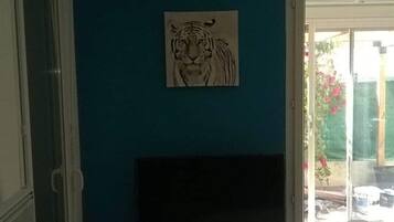 Ein Zimmer mit blauer Wand, einem aufgehängten Tigerbild, einem Fernseher auf einem weißen Standfuß und einer Glastür, die nach draußen führt.