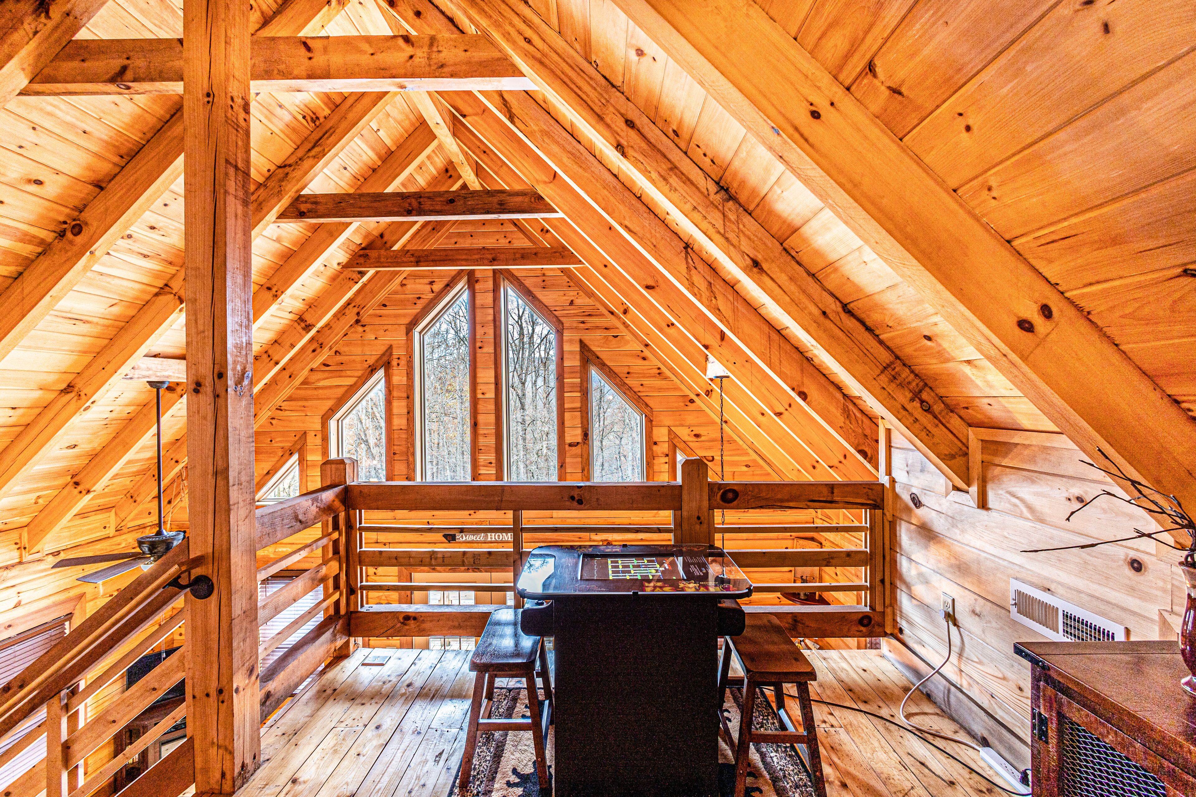A wooden cabin with a loft bedroom featuring a desk and two chairs, large windows, and a wooden railing.