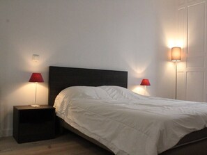 Cama de solteiro com cabeceira escura em quarto com paredes brancas. Há duas mesas de cabeceira, cada uma com um abajur vermelho.