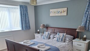 Un dormitorio ordenado con una cama de madera, una colcha de rayas azules y blancas, dos mesitas de noche con lámparas, una ventana con cortinas azules y una obra de arte colgada en la pared.