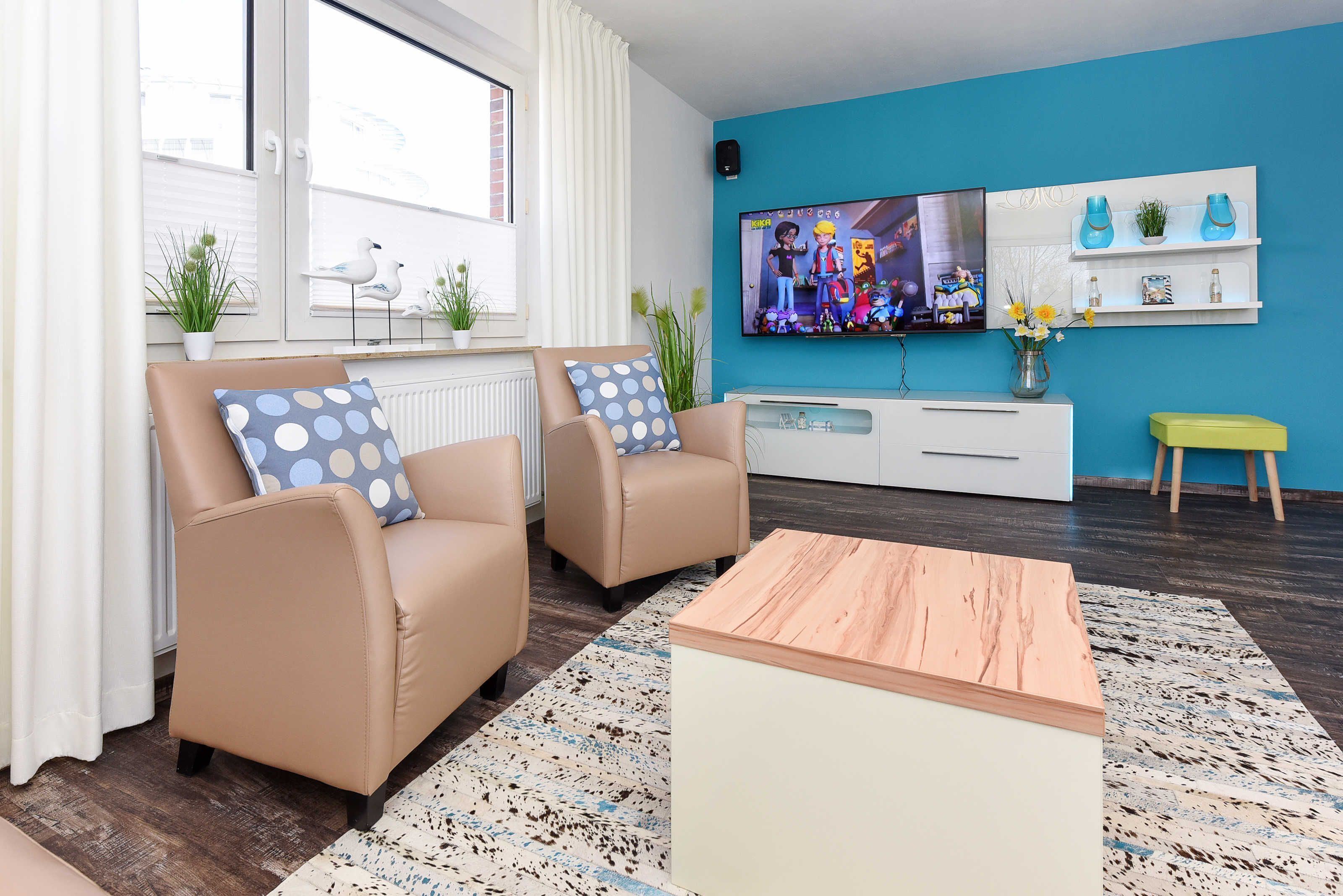 Un salon moderne avec deux fauteuils beiges, une table basse en bois, un téléviseur fixé au mur et un meuble blanc agrémenté d’objets décoratifs.