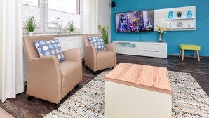Un salon moderne avec deux fauteuils beiges, une table basse en bois, un téléviseur fixé au mur et un meuble blanc agrémenté d’objets décoratifs.