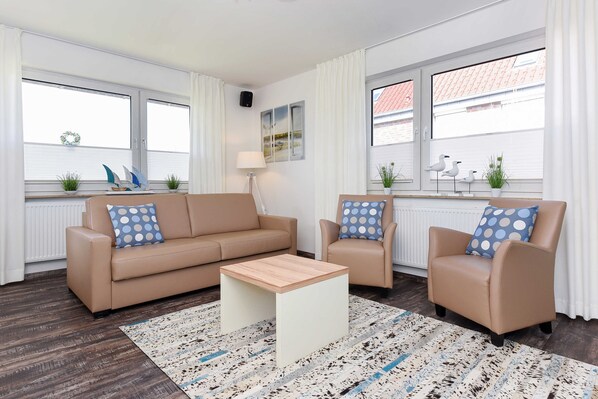 Una sala de estar moderna con un sofá color beige, un sillón a juego y una mesita de centro. Hay dos ventanas con cortinas, un radiador y objetos decorativos en los alféizares.