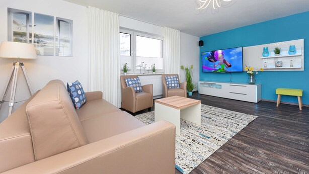 Un salon moderne avec un canapé beige, deux fauteuils, une table basse, un téléviseur et un mur d’accent bleu.
