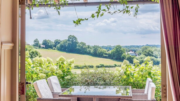 A wicker table and chairs with a glass top, set against a backdrop of a lush green landscape and a clear sky. - Castlemans Stables East (Sedlescombe, near Battle)