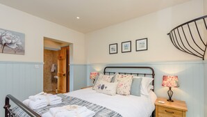 A neatly made bed with a patterned bedspread, a bedside table with a lamp, and a wooden door leading to another room. - Castlemans Stables East (Sedlescombe, near Battle)